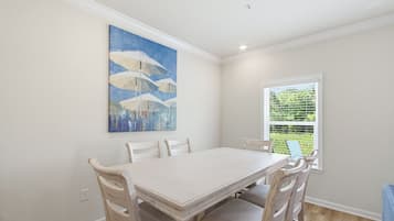 A dining room with a white table and chairs, a painting on the wall, and a window with blinds.