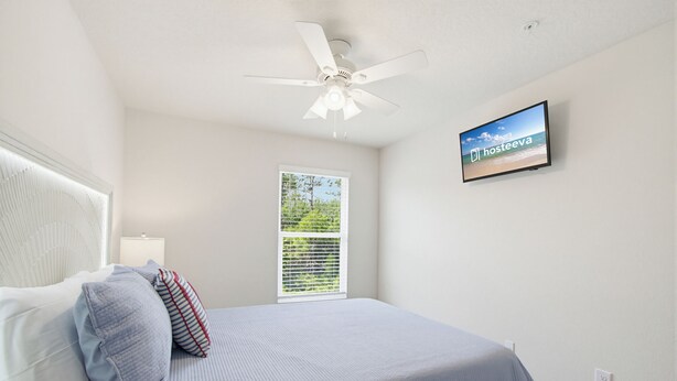 Un dormitorio con cama, almohadas, ventilador de techo, televisión y ventana con persianas.