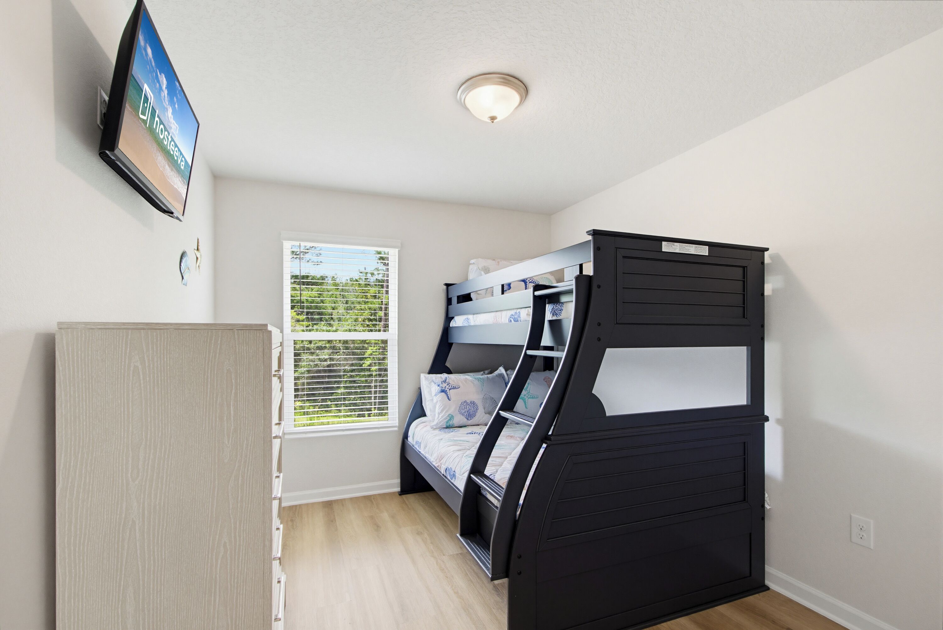 A bunk bed room with a television on the wall, a window with blinds, and a ceiling light fixture.