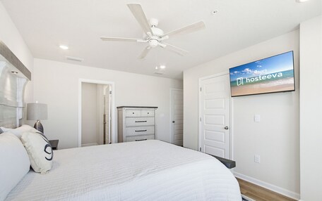 A bedroom with a bed, a ceiling fan, a TV on the wall, and a dresser.