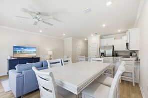 A modern living room with a sofa, dining table, chairs, a TV, ceiling fan, and kitchen area. - Lost Bay Hideaway (Orange Beach)