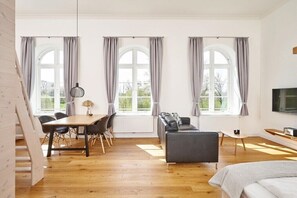 A spacious room with a wooden floor, a dining table with chairs, a sofa, a bed, and large windows with curtains. - Holiday apartment with 1 bedroom (Steinberg)