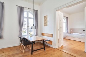 A room with a wooden floor, a dining table with chairs, a bench, a bed, and a window with curtains. - Holiday apartment with 1 bedroom (Steinberg)