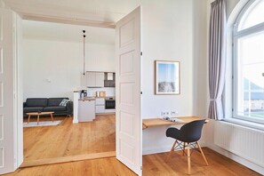 A room with a wooden floor, a black chair, a desk, a window with curtains, and a kitchen area visible through an open door. - Holiday apartment with 1 bedroom (Steinberg)
