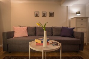 A living room with a grey sofa, a white side table, a vase with flowers, books, and a lamp. - Holiday apartment with 2 bedrooms (Naumburg)