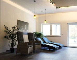 Ein Zimmer mit zwei Korbstühlen, einem Liegestuhl, einer Zimmerpflanze, einem gerahmten Bild an der Wand und einem großen Fenster.