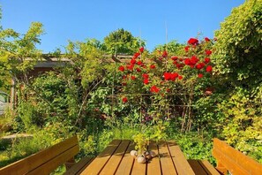 Eine hölzerne Bank mit Tisch in einem Garten, der von einer Kletterpflanze und roten Blüten geschmückt ist.
