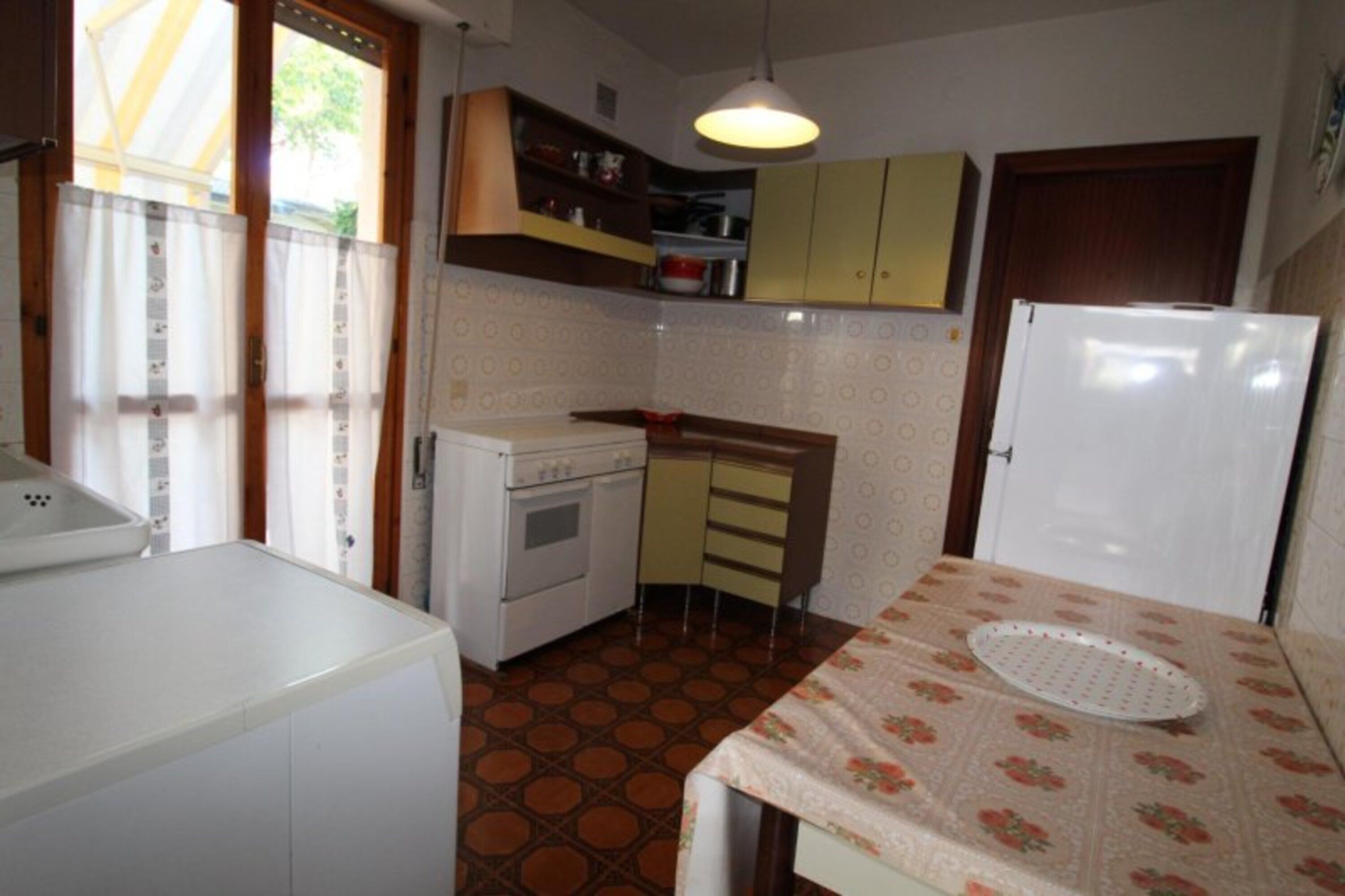 Uma cozinha compacta com fogão, forno e geladeira, uma mesa de jantar pequena e uma janela com cortinas.