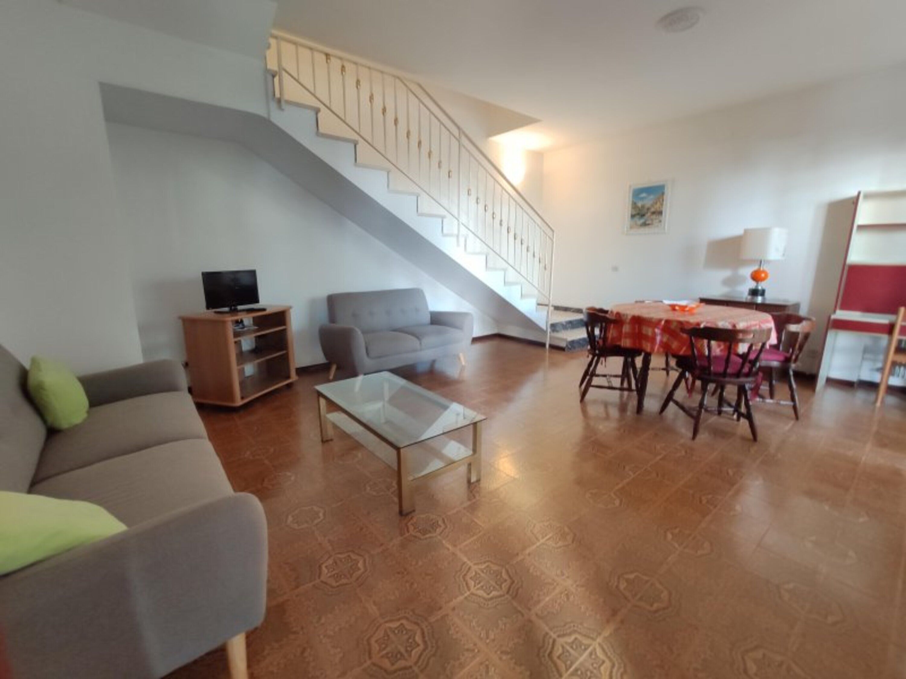 A living room with a sofa, a coffee table, a TV stand, a staircase, and a dining area with a table and chairs.