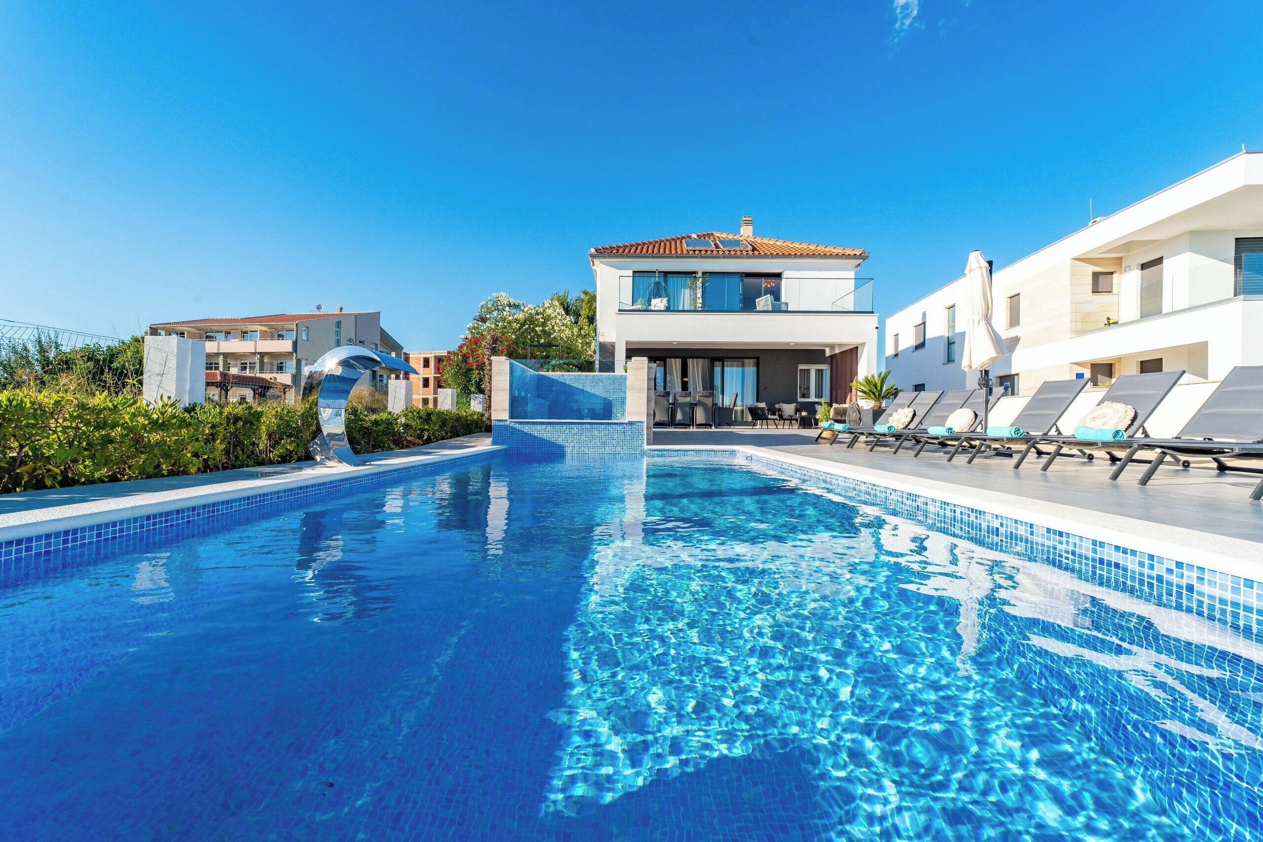 A swimming pool with clear blue water, surrounded by modern buildings and lounge chairs.