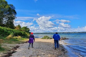 Zwei Personen spazieren am sandigen Strand entlang, im Hintergrund Wasser und Bäume.
