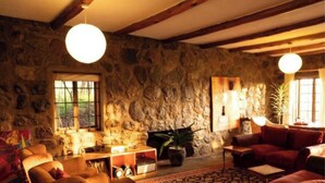 A rustic living room with stone walls, wooden beams, a fireplace, and a large window. - Oloile - Home By The Park (Nairobi)