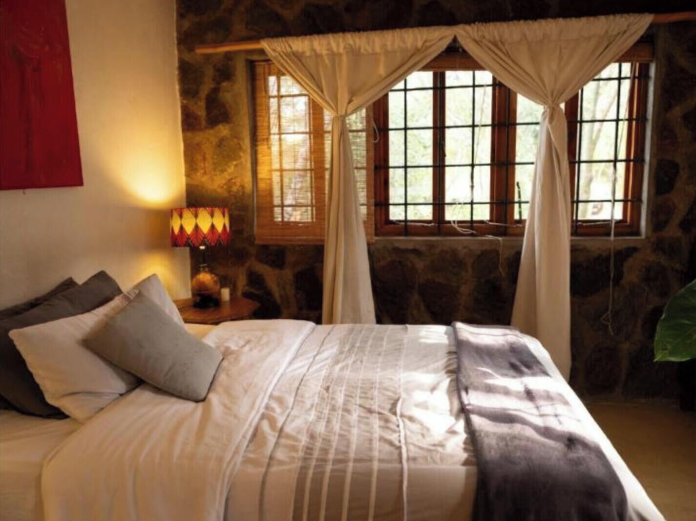 Une pièce comprenant un lit, une table de chevet avec une lampe, un mur en pierre et une fenêtre avec des rideaux.