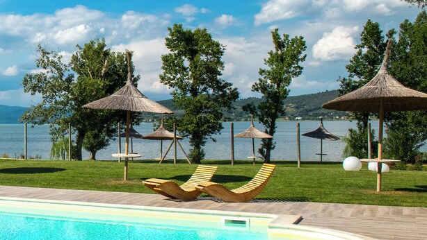 A swimming pool with lounge chairs and thatched umbrellas, surrounded by trees and a view of a body of water. - Villa with pool and garden on Lake Trasimeno (Magione)