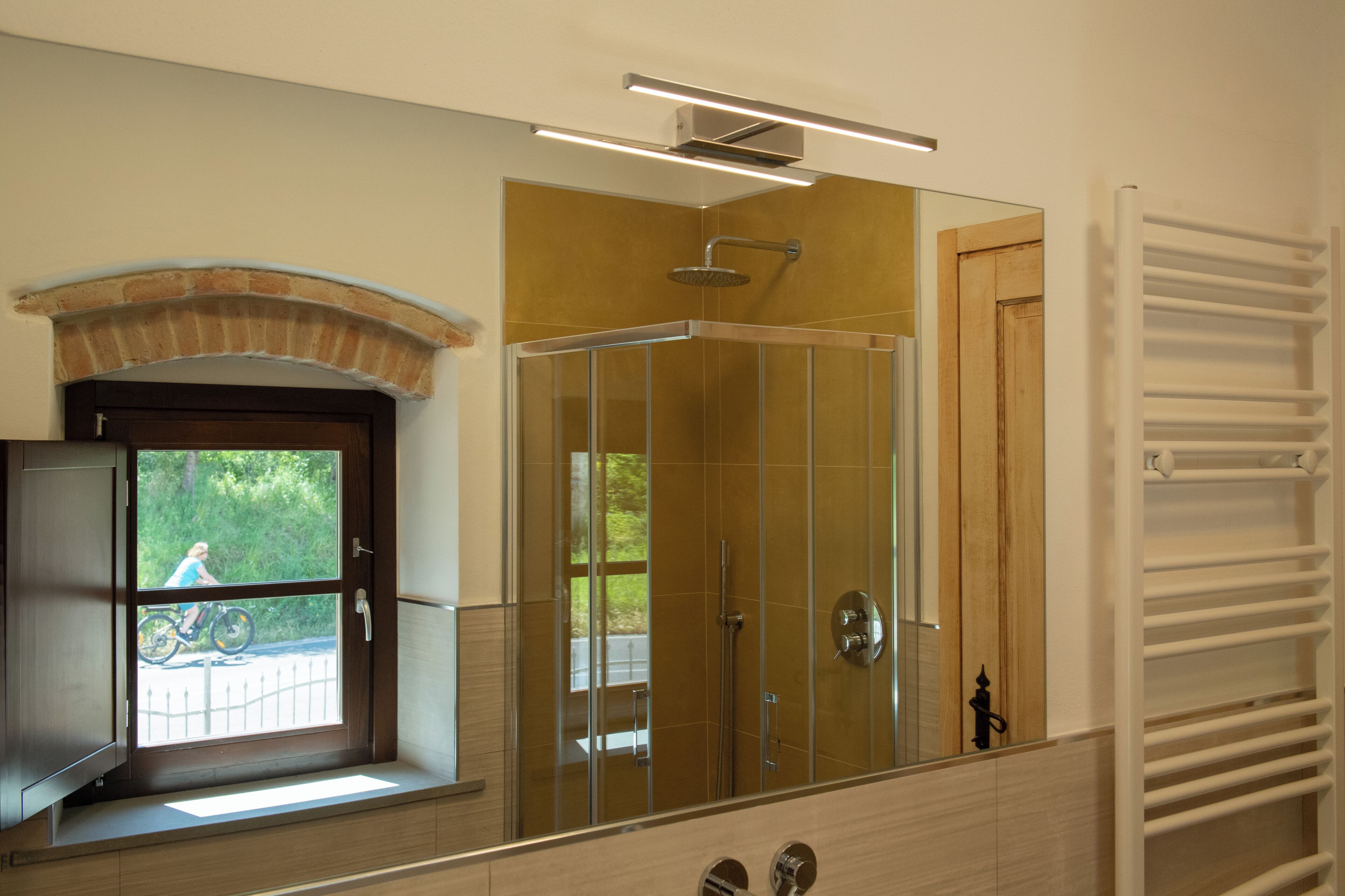 Une salle de bain avec une douche à parois de verre, une fenêtre donnant sur une personne à vélo et un porte-serviettes blanc.