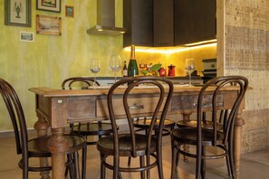 A dining area with a wooden table and chairs, a kitchen in the background, and framed artwork on the wall.