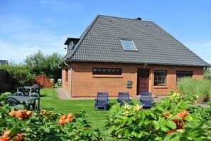 Ein Haus mit geneigtem Dach, einem kleinen Fenster und einer Ziegelwand. Zwei blaue Stühle stehen draußen, und im Vordergrund befindet sich ein Tisch mit Stühlen.