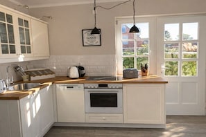 Eine Küche mit weißen Schränken, einem Geschirrspüler, einem Backofen, einer Kaffeemaschine und einem Fenster mit Blick ins Grüne.