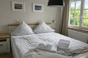 Ein ordentlich gemachtes Bett mit weißen Leinen und Kissen, ein Nachttisch mit Lampe und ein Fenster mit Blick auf Bäume.