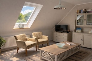 A loft-style room with a sloped ceiling, a television, two armchairs, a wooden table, a bookshelf, and a window with a view of greenery. - Oland in the house Achter de Dünen (Sankt Peter-Ording)