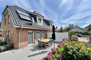 Ein Wohnhaus mit Schrägdach, Solaranlage, Ziegelaußenwand, einer Terrasse mit Tisch und Stühlen sowie einem Blumenbeet.