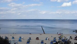 Ein Strand mit blauen Liegestühlen und Sonnenschirmen, einer Pier, die ins Wasser ragt, und einem klaren Himmel.