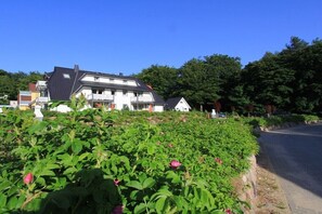 Ein Wohngebiet mit einem zweigeschossigen Haus, einem Garten mit grünem Laub und rosa Blüten, sowie einer Straße im Hintergrund.