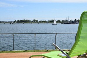Eine Holzterrasse mit einem grünen Stuhl, Blick auf ruhiges Wasser und einem Segelboot in der Ferne.