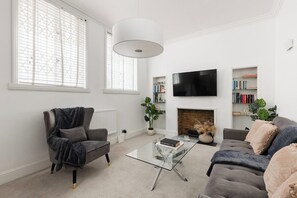 Ein Wohnzimmer mit Sofa, Sessel, Glastisch, Kamin, Fernseher und Bücherregalen.