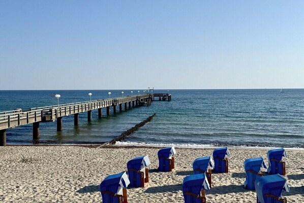 Ein Strand mit einem Steg, der ins Meer führt, und mehreren blauen Stühlen, die am Sand aufgereiht sind.