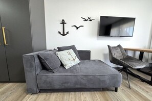 Un salon moderne avec un canapé d’angle gris, un fauteuil gris, un téléviseur à écran plat et des éléments de décoration murale, notamment une ancre et des oiseaux.