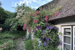 A rustic building with a thatched roof, surrounded by a garden with various flowering plants. - "The Coachman's Cottage" (Langballig)