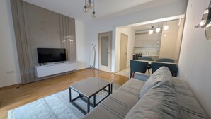 A modern living room with a flat-screen TV, a sofa, a coffee table, and a kitchen in the background. - Status M Apartments 98 (Becici)