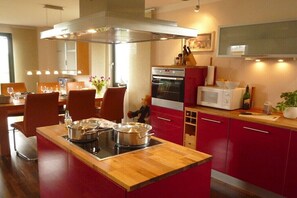 A modern kitchen with red cabinets, a central island with a stove, and a dining area with chairs and a table. - Thatched House Four Seasons (Insel Poel)
