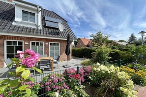 Ein Haus mit Ziegelverkleidung, einem Ziegeldach, einer Terrasse mit Gartenmöbeln und einem Garten mit verschiedenen Blumen.