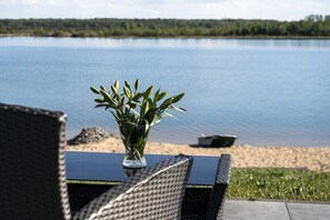 Ein Tisch mit einer Glasvase voller Grünpflanzen, mit Blick auf einen ruhigen See und einen sandigen Uferstreifen mit Bäumen im Hintergrund.