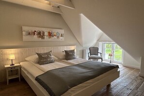 A bedroom with a large bed, a nightstand, a chair, a window, and a painting on the wall. - "Reethus" in the house on the dike (Vollerwiek)