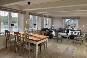 A spacious room with a dining table, chairs, a sofa, and a coffee table. There are large windows with curtains, and the room has a high ceiling. - "Reethus" in the house on the dike (Vollerwiek)