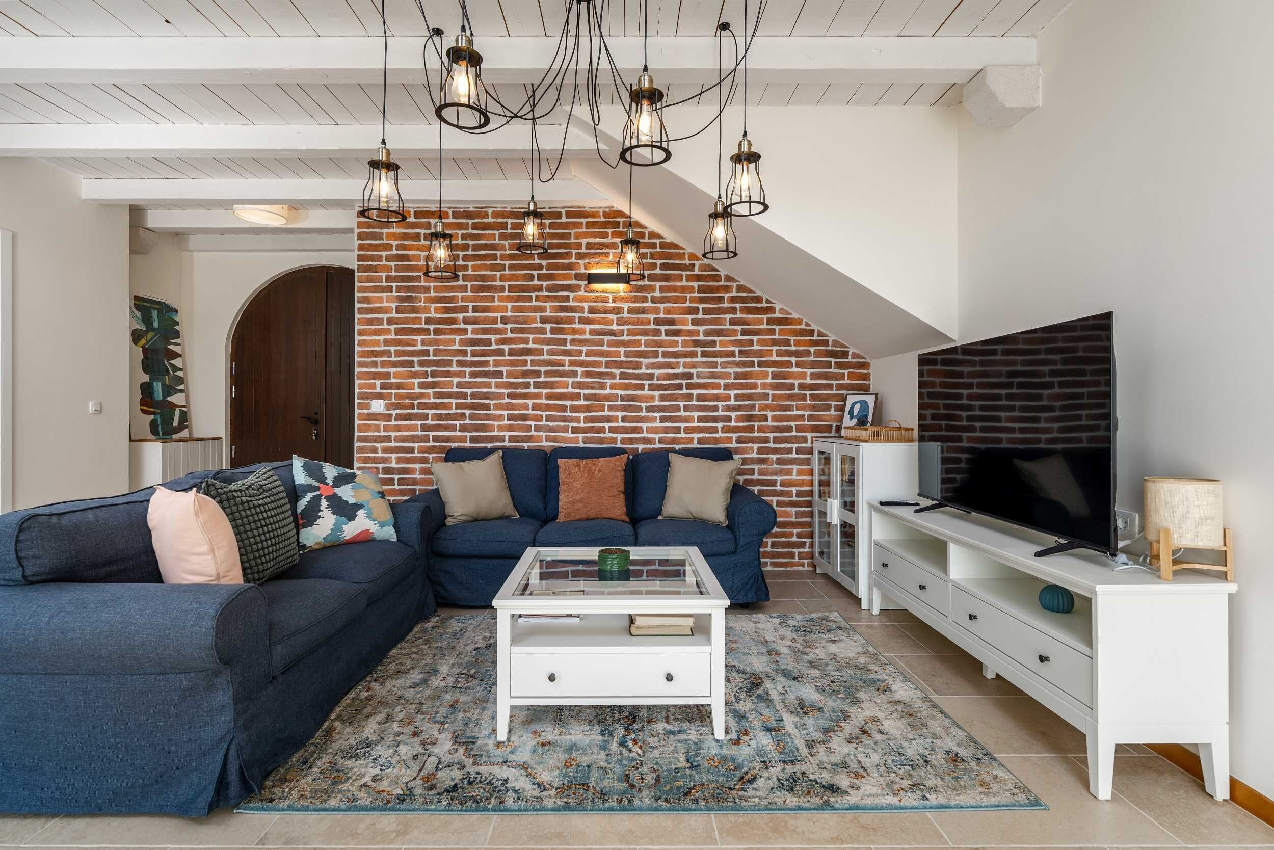 Un salon avec un mur en briques, un canapé, une table basse, un téléviseur et des luminaires suspendus.