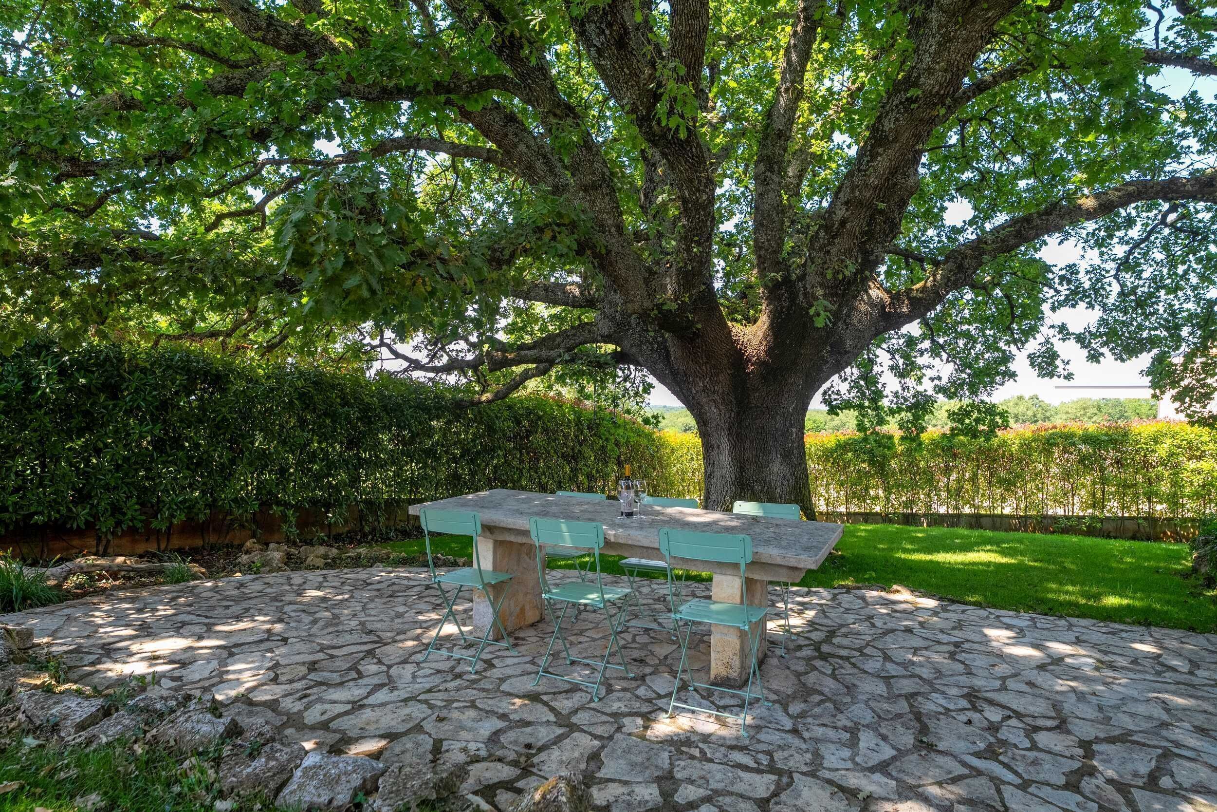 A stone patio with a table and chairs under a large tree.