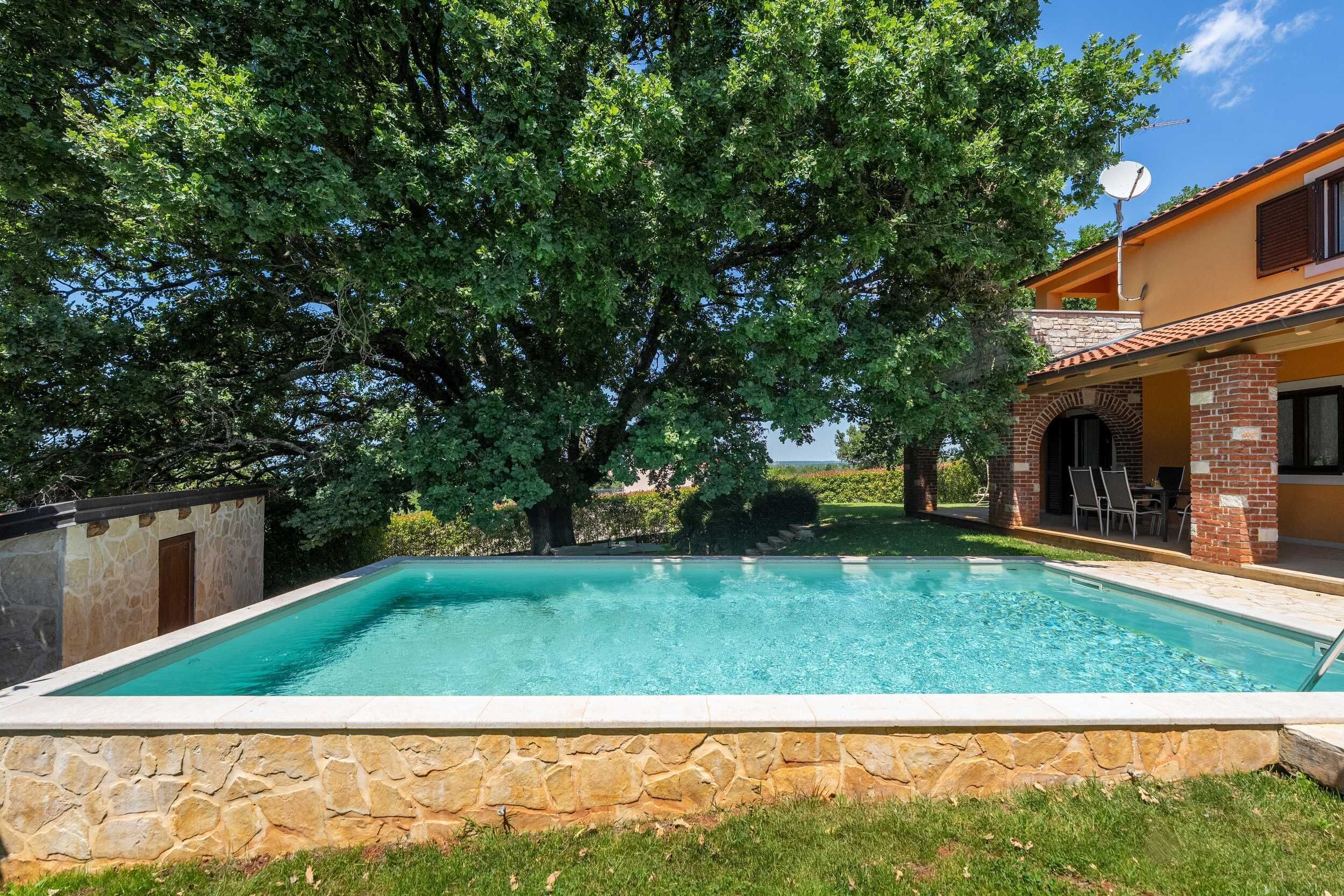 Una piscina rectangular con paredes de piedra, rodeada de césped y árboles, con un edificio con tejado de azulejos y ladrillo en el fondo.