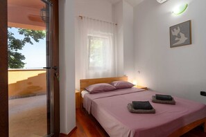 Ein Schlafzimmer mit einem Bett, einem Fenster mit transparenten Vorhängen, einem Nachttisch und einer Wandleuchte.