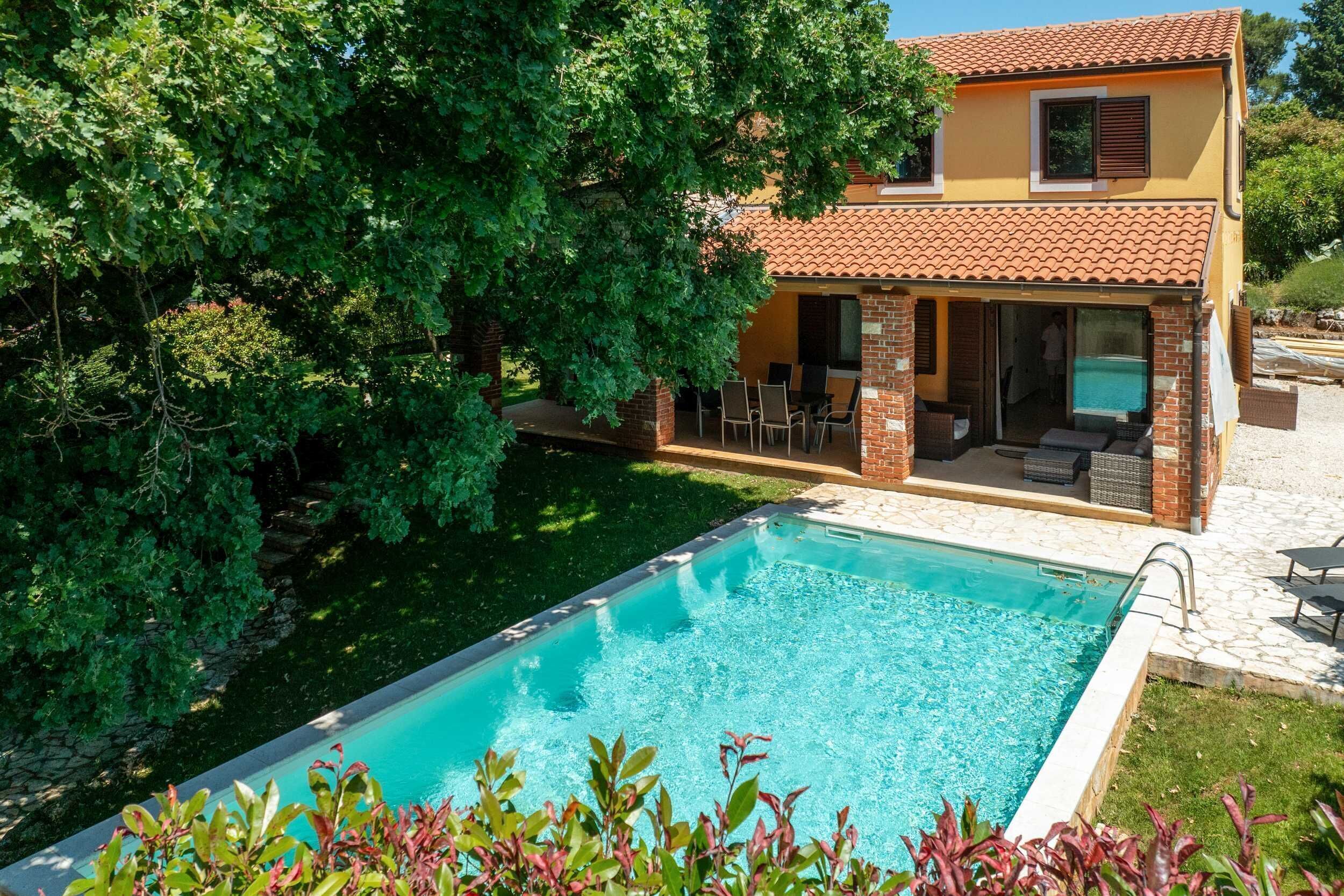 Una piscina con zona de patio, un edificio de dos plantas con tejado de tejas y vegetación circundante.