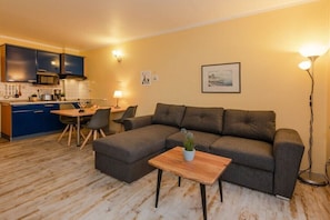Ein Wohnzimmer mit einer Couch, einem kleinen Tisch mit einer Pflanze, einem Essbereich mit Stühlen und einer Küche im Hintergrund.