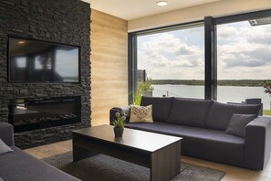Ein modernes Wohnzimmer mit Kamin aus Stein, einem großen Fernseher, einer Couch und Blick auf einen See.
