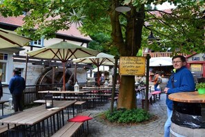 Ein Straßencafé mit Holztischen und -bänken, Sonnenschirmen und einem Steinhaus im Hintergrund.