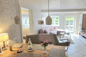 A dining area with a wooden table, a vase with flowers, a bottle of Veel, and hanging pendant lights. - Holiday apartment with 1 bedroom (Oldsum)