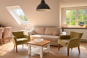 Ein gemütliches Wohnzimmer mit einer Couch, zwei Sesseln, einem kleinen Tisch, einer Pendelleuchte und einem Fenster mit Blick ins Grüne.