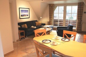 Un salon chaleureux avec un canapé, une table à manger dressée pour deux personnes et une grande fenêtre agrémentée de rideaux.