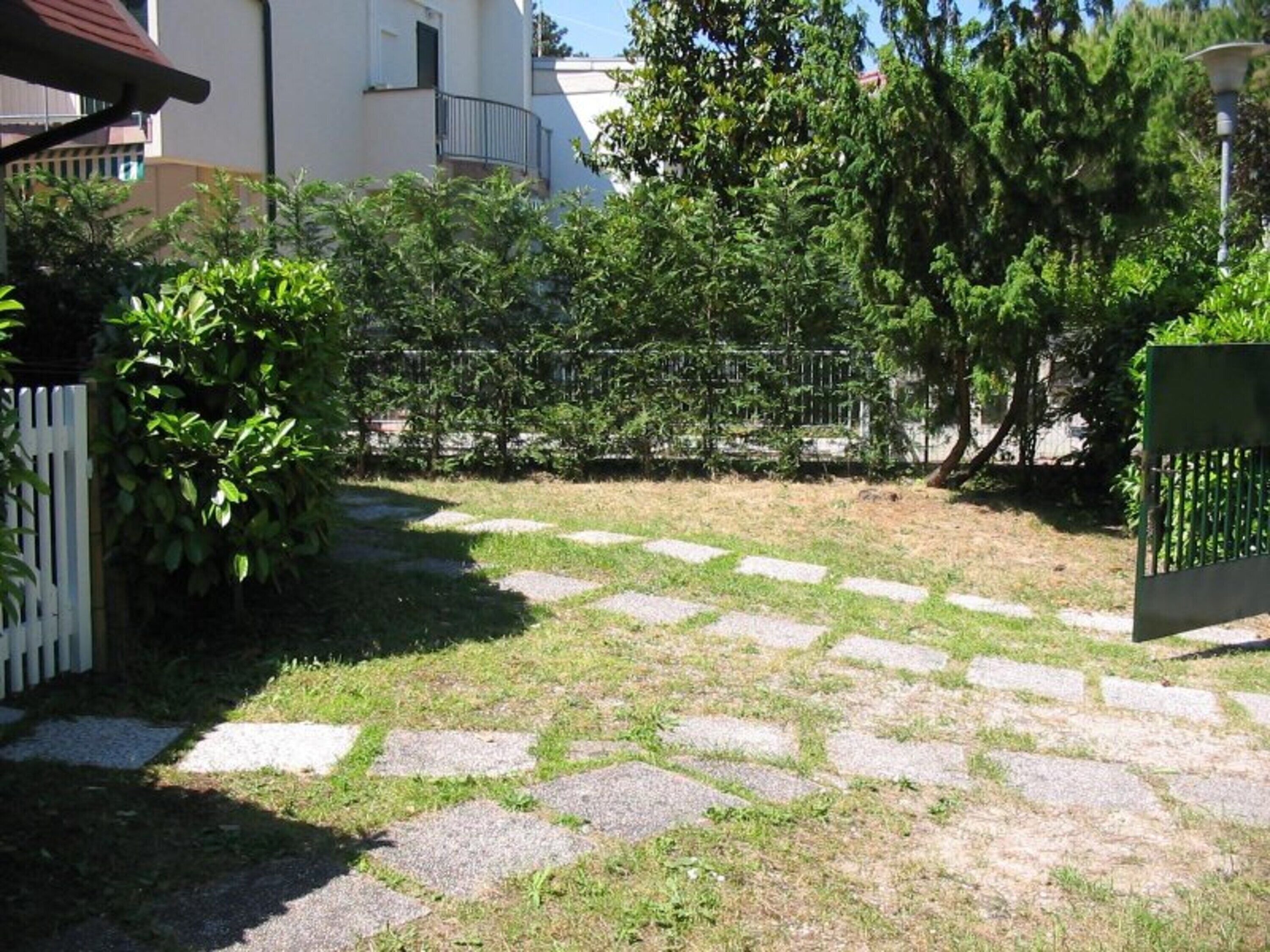 A paved courtyard with a white fence, greenery, and a building in the background.